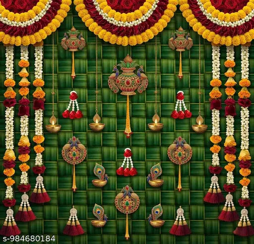 A festive decorative display featuring a green woven bamboo or palm leaf backdrop adorned with yellow and maroon flower garlands, hanging brass oil lamps, ornate wall ornaments, and peacock and floral motifs.